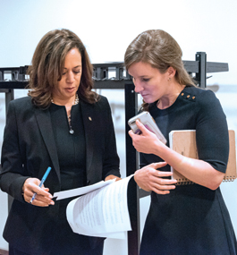 Kamala Harris and Lily Adams, both wearing dark clothes, look down at papers Harris is holding. Adams holds a cell phone and a notebook.