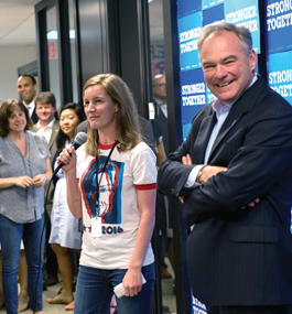 Lily Adams, wearing a Hillary Clinton 2016 T-shirt and jeans, stands next to Tim Kaine in a room with "Stronger Together" posters on one wall. Lily holds a microphone; campaign workers watch and smile.
