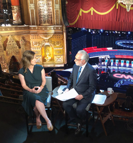 Up in a balcony in an ornate theater, with podiums and the CNN logo visible on the stage below, Lily Adams talks with Wolf Blitzer (both seated).