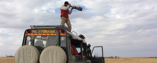 On a grassy plain against a blue sky with white and gray clouds, a man looking through a camera with a large lens stands on the roof of a jeep-like vehicle.