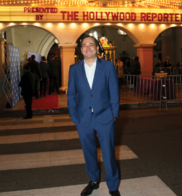 A smling man in a blue suit stands in a crosswalk outside a building fronted by a fancy theater marquee that shows the words "Presented by The Hollywood Reporter."