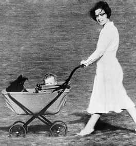 A dark-haired woman in a white dress pushes a baby carriage outside; peeking over the side of the stroller is a black dog and a curly-haired baby.