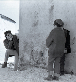 A young man peers around a roughly plastered wall to take a photo of two children
