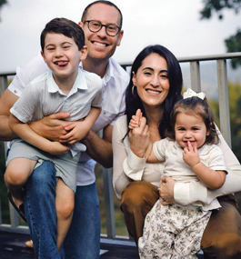 A man wearing glasses holds a young boy who has his tongue out in a smile, and a woman holds a smiling young girl with a white bow in her hair.