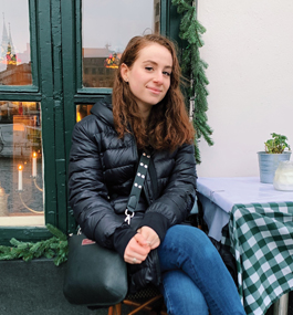 Woman in dark puffer jacket and jeans with a leather satchel slung over her shoulder sits at an outdoor table with a green checkered covering.