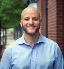 Head shot of a smiling man with a beard wearing a light-blue shirt, taken outside near some trees and a brick building.