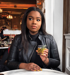 A young woman wearing a black leather jacket, looking slightly away from the camera, sits at a table with a bar in the background holding up a cocktail.