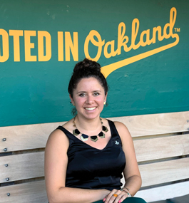 A smiling young woman sits on a bench in front of a green wall that has "Rooted in Oakland" painted in yellow letters.