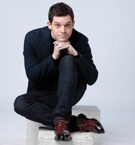 A man wearing dark clothes is seating in front of a white background with his chin and hands resting on one knee.