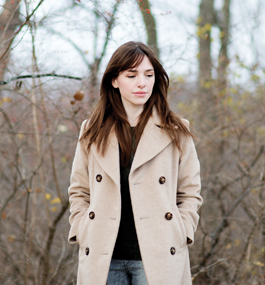 A woman stands in a late-autumn woodland setting with her hands in the pockets of her beige coat.
