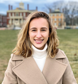 A young woman in a beige wool coat stands outside with stately mansions in the background.