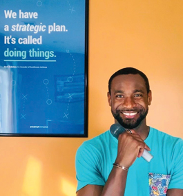 A smiling man holding a microphone in one hand stands in front of a poster that reads, in part, "We have a strategic plan. It's called doing things."