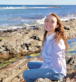 A young woman in jeans sits on a rocky coastline, smiling broadly for the camera.