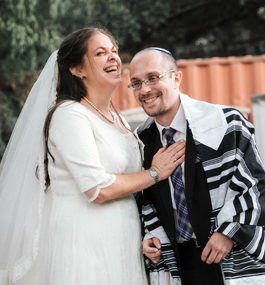 A woman in a white dress and veil and a man wearing a prayer shawl, both smiling, lean toward each other.