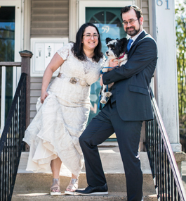 A man holding a small back-and-white dog and a woman in a white dress pose on steps leading up to a house.