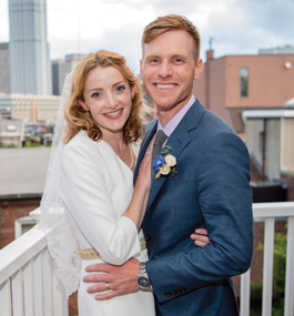 A smiling, embracing man and woman on a roof deck that overlooks a Boston skyline.