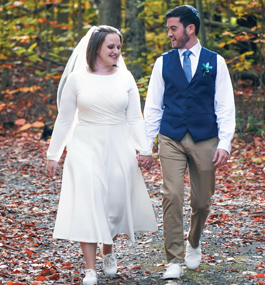 A woman in a mid-calf white dress and veil and a man in a blue vest, white shirt and khakis — both wearing white sneakers — hold hands and walk through a woodland setting.