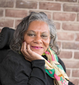 Photo of a woman with gray hair wearing a brightly colored scarf posing with her chin resting on her hand in front of a brick wall.