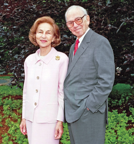 Photo of a man in a gray suit and red tie standing with a woman in a pale-pink suit in front of green foliage.
