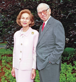 Carl Shapiro with his wife, Ruth Photo of a man in a gray suit and red tie standing with a woman in a pale-pink suit in front of green foliage.