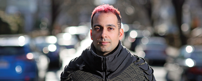 Portrait of a man whose dark hair has pink tips, standing outside in a heavy jacket in hazy winter light.