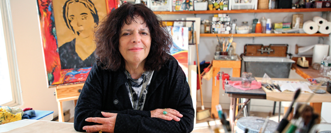 A woman with dark curly hair sits at a table with colorful art behind her.