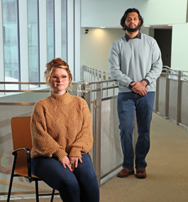 A seated woman in a brown sweater and standing man in a light-gray sweater in a light-filled interior space.