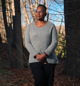 A woman in gray sweater standing outside under bare trees looks off into the distance.