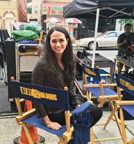 A dark-haired woman sits in a navy-blue director's chair with a "Blue Bloods" logo on the back.