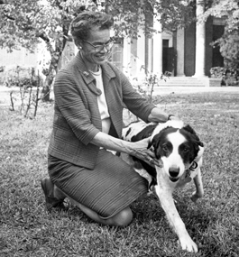 A black-and-white of a smiling woman kneeling on grass, holding a dog