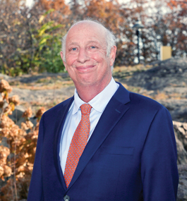 Head shot of a smiling gray-haired man in a dark-blue suit and reddish tie