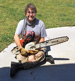 Women wearing heavy work gloves sits on the ground outside, holding a chain saw in her lap