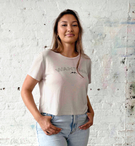 A long-haired woman in a T-shirt stands in front of a rough brick wall painted with white paint