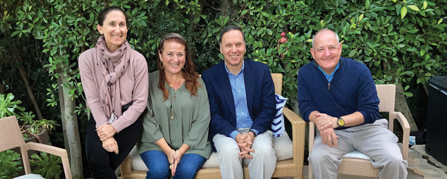 Four smiling people sit on a patio bench and chair against a background of greenery