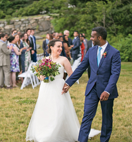 A bride and groom, smiling broadly at each other, walk while holding hands