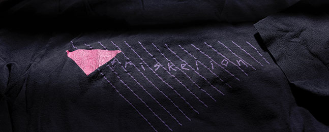 A shadowy photo of a dark T shirt emblazoned with a pink triangle and the name "Triskelion."