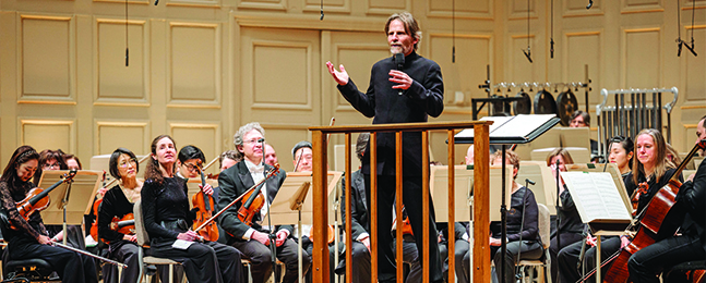 A man speaks at a podium with formally dressed orchestra members behind him.