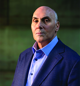 Headshot of an unsmiling man wearing an open-collar shirt and dark-blue coat.