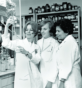 Three women in white lab coats look at a beaker in a laboratory