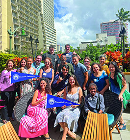 A group of people, some holding Brandeis banners, smile for the camera in an tropical outdoor setting