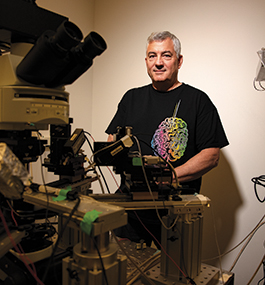 Photo portrait of Sacha Nelson, wearing a T-shirt that shows an image of the human brain, standing behind lab equipment.