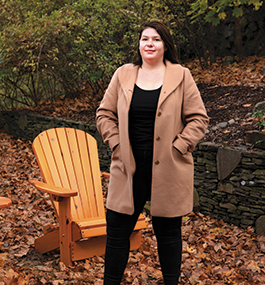 Photo portrait of Anne Berry, wearing a light-brown coat, standing outside in front of an Adirondack chair.