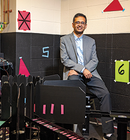 Photo portrait of Shantanu Jadhav, wearing a gray blazer and a button-down shirt, seated in front of lab equipment.