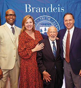 President Ron Liebowitz and three other people smile for the camera in front of a blue Brandeis banner.