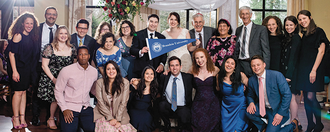 A bride and groom holding a Brandeis pennant are surrounded by smiling wedding guests.