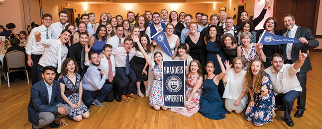 A large group of people pose for the camera behind a Brandeis banner.
