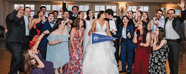 A bride holding a Brandeis pennant kisses a groom while surrounded by many cheering people.