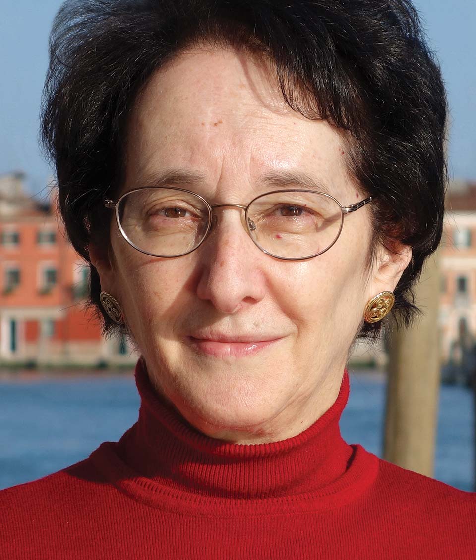 Naomi Baron wearing wire-frame glasses, gold earrings and a red turtle-neck sweater.