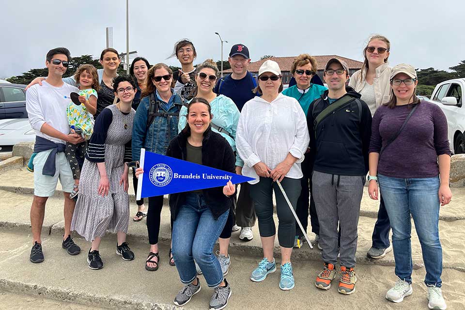 A group of Brandeis alumni pose on the beach. The person in front holds a Brandeis pennant.