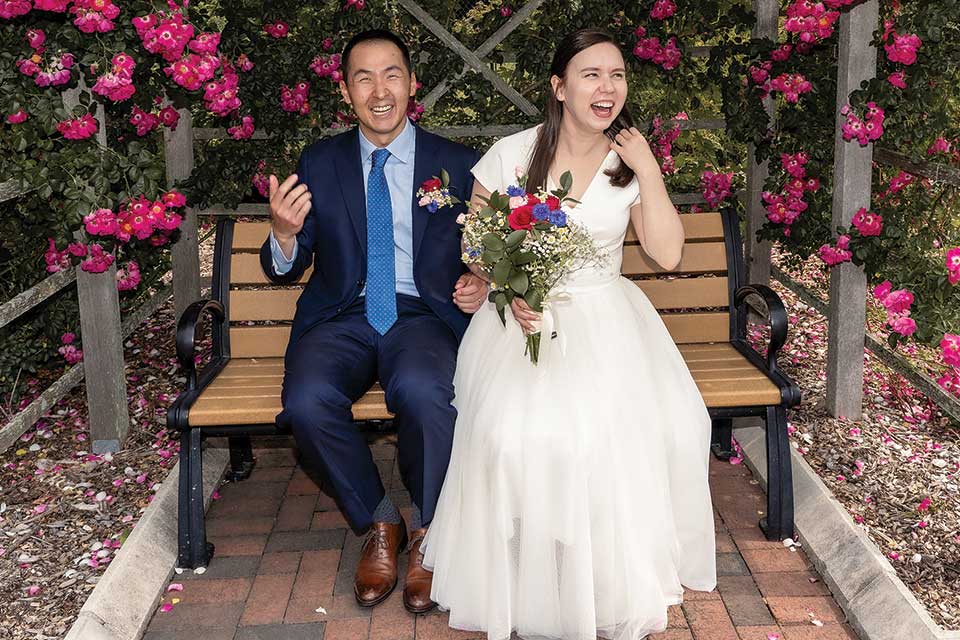 A bride and groom laugh while sitting on a bench surrounded by flowers